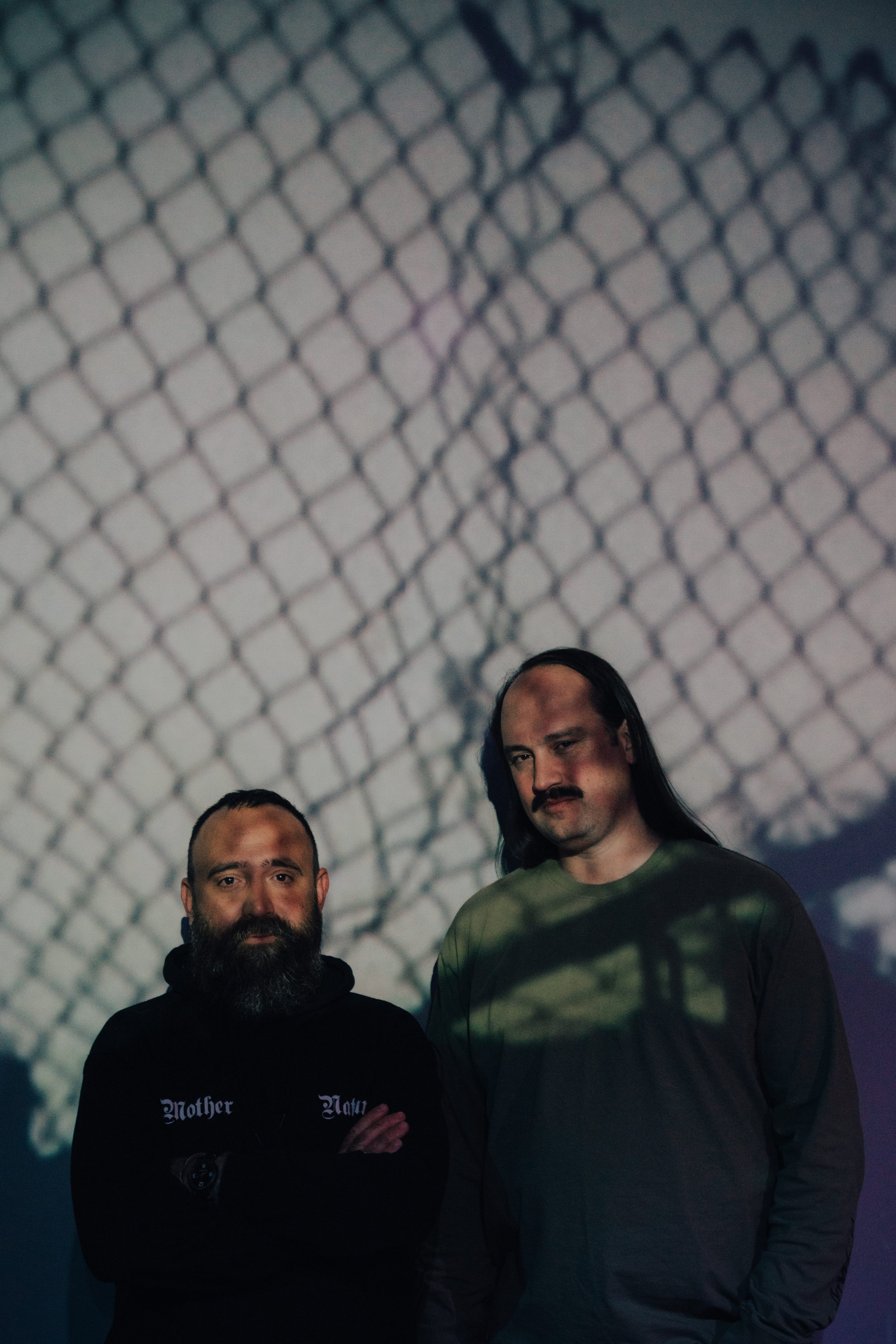 Pete Swanson and Gabriel Saloman Mindel stand side by side, a chainlink fence projected over their faces, it's dark and moody looking.
